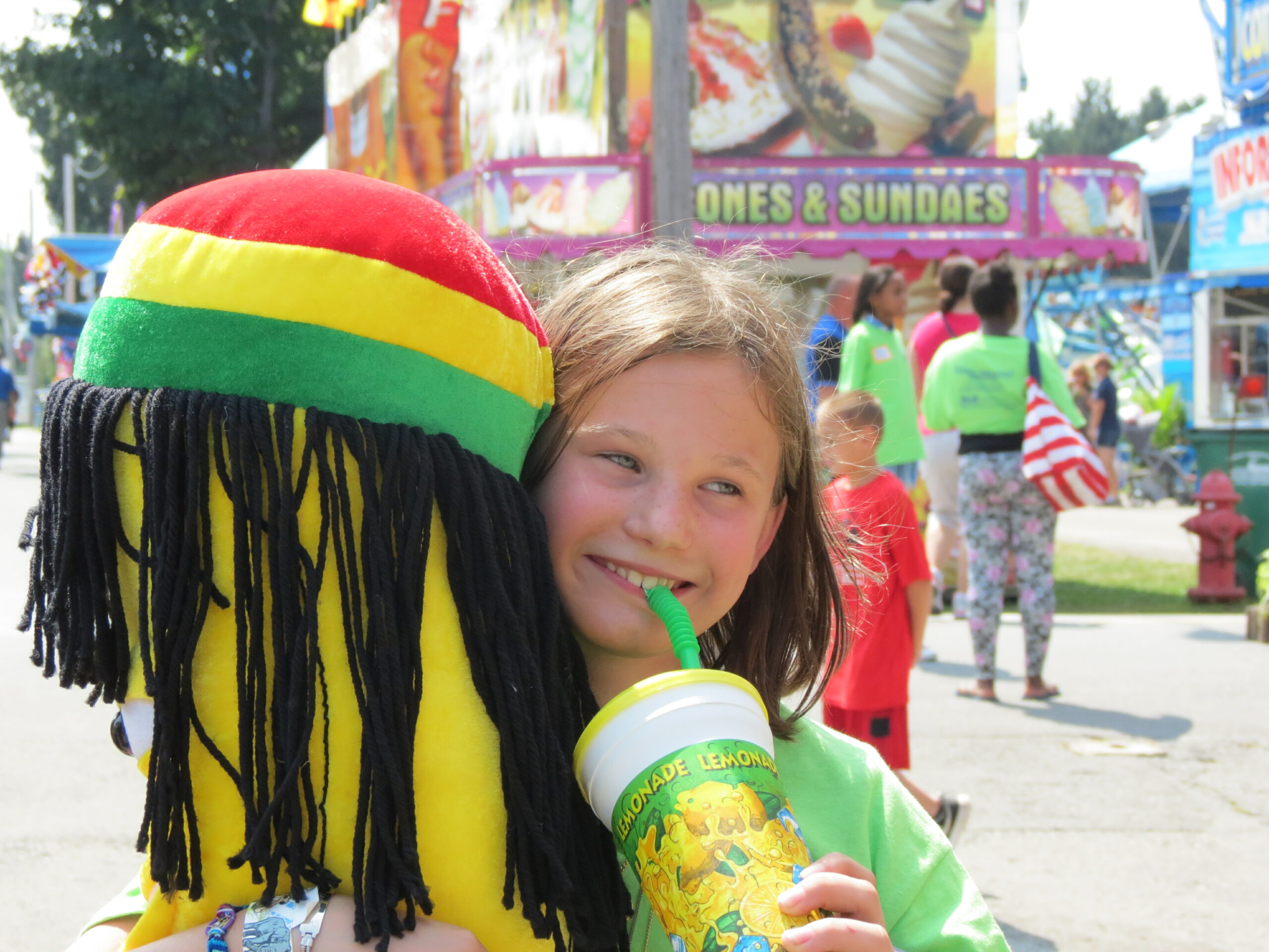 A young child holds a stuffed prize and sips a lemonade at a fairground. In kind donations to Astor Services make such joy possible.