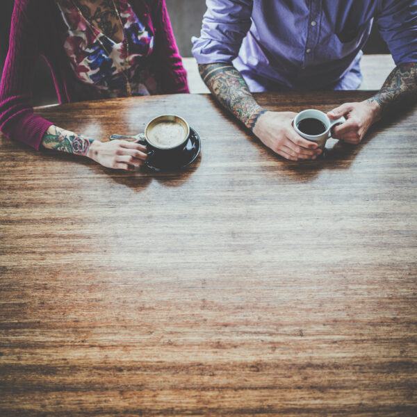 two people sit at a rustic wooden table with hot coffee and espresso in mugs. The people are dressed business casual and have tattooed arms. Their faces are out of frame. Practice compassion