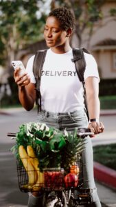 A Black person wearing a backpack and a t-shirt that reads, "Delivery" stands with their bicycle loaded with groceries and reads their phone for directions. Great idea for a grocery meal train