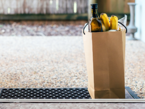 a paper bag full of groceries like olive oil and bananas sits on a welcome mat in front of a home's open front door. Grocery delivery is a great part of a no-contact meal train or no-contact food drop off