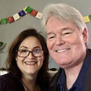 Posing for a self-portrait, Melinda a woman with dark brown, straight, long hair and dark rectangular glasses, and Kyle -- a man with short grey hair smile at the camera together
