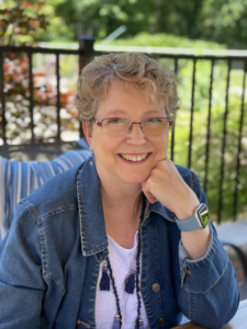 Pamela, a denim jacket clad, short blonde curly haired woman in glasses, is seated outdoors, smiling warmly, resting her chin on her left hand, which wears an apple watch.