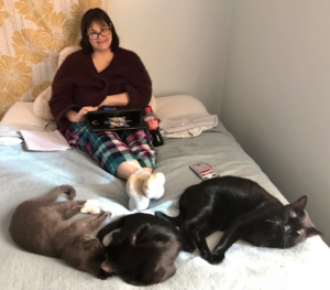 On a bed, three cats are casually laying on their sides at the feet of a woman seated, feet crossed over one another, wearing glasses and smiling at the camera.