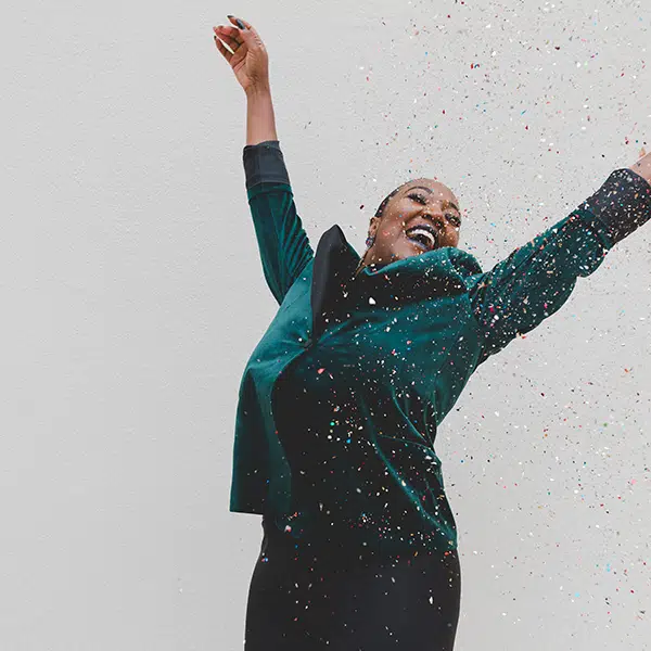 Black woman wearing an emerald green, velvet suit, raises her arms in celebration amid a shower of glitter confetti. gifts for friend with new job