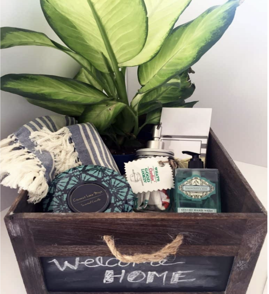 rustic, wooden gift box filled with a potted plant, blanket, hand soap, scented candle, and chocolate. the outside of the box is chalkboard material and displays the chalk writing, "Welcome Home". what to get someone with a new house