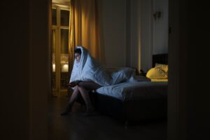 person with a blanket wrapped around their head and body sits in a dark room illuminated by the glow of a television