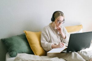 person sitting in bed with a laptop computer, writing in a notebook, and wearing glasses and headphones