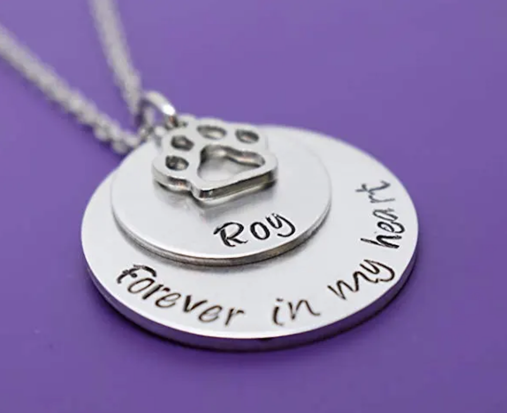 what to get for someone who lost a pet: custom stamped, round, silver medallion hanging from a necklace reading "Roy, forever in my heart" and a paw-print shaped silver charm stacked on top