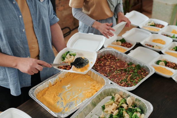 foil pans of macaroni and cheese, beans, and broccoli & cauliflower are on a table. People scoop servings into to-go containers. recipes for meal train