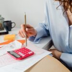 A woman sits at a table among a scattering of paper bills. She uses the back of a pencil to type into a calculator.