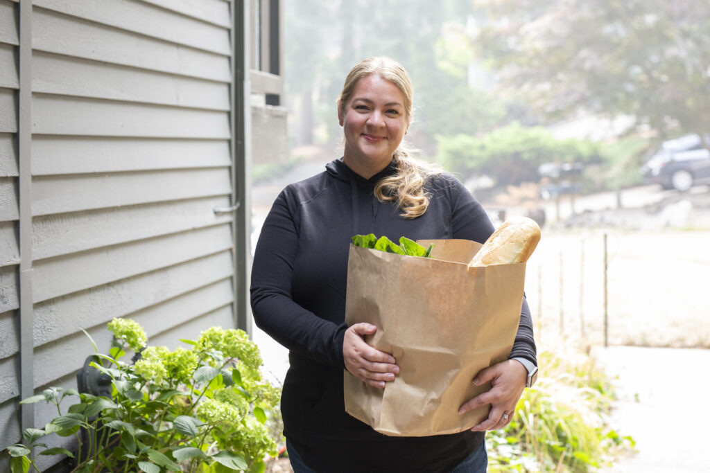 Smiling woman holding a bag of groceries outside a friend's home.