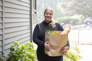 Smiling woman holding a bag of groceries outside a friend's home. Delivering groceries is an innovative answer to the question, What is a meal train?