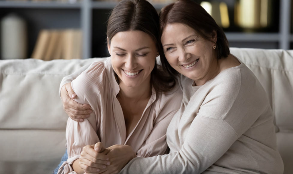 Two smiling women sit side by side on a couch, one wraps the other in an embrace.