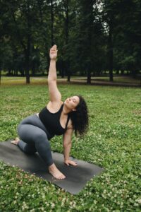 A woman practices yoga in a park on a yoga mat. She kneels on her mat reaching and looking up toward the sky.