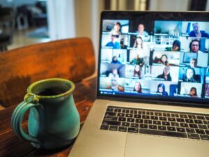 A laptop computer displaying a gallery view of people on a zoom call sits next to a coffee mug on a table.