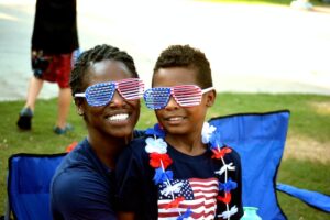 Outside, sitting in a picnic chair, a Black mother and son wear American flag style sunglasses, he also wears an American flag t-shirt and red, white, and blue lei around his neck. They smile at the camera.
