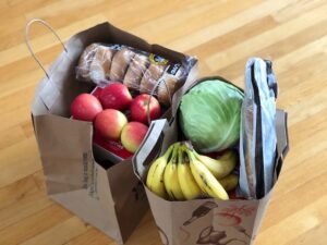 Two paper grocery bags sit on a hardwood floor. They are filled with fresh items like apples, bananas, lettuce, and bagels