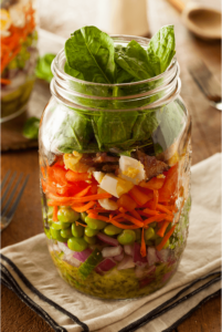 a mason jar is filled with layers of salad dressing, legumes, herbs, boiled eggs, and leafy greens. mason jar salads are great summer meal train ideas