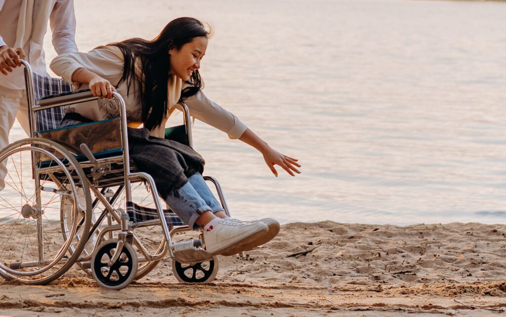person in wheelchair at the beach reaching down to the sand and smiling