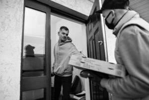 Black and white image of a man answering the door to a pizza driver wearing a baseball cap and a face mask and holding 2 pizzas.