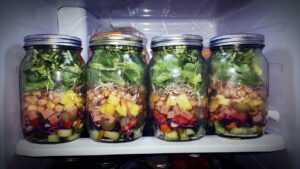 Mason Jars full of layered salad lined up on a refrigerator shelf, great summer meal train ideas