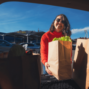a smiling woman places a paper bag full of groceries into the trunk of her car. the sun brightly shines on her face and the grocery bag.