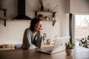 woman stands in kitchen at a counter smiling at a laptop computer. She appears to be on a video call. Video calls are a welcome visit during post operative care