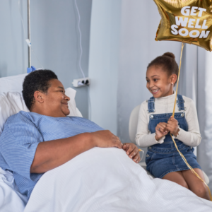 a young person sits in the hospital next to a patient in a hospital bed. The young person is holding a gold balloon that reads "get well soon"
