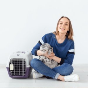 a person sits on the floor with their legs crossed under them. They hold a cat in their lap and they both sit beside a hardshell pet carrier. planning for pet care during emergency is so important